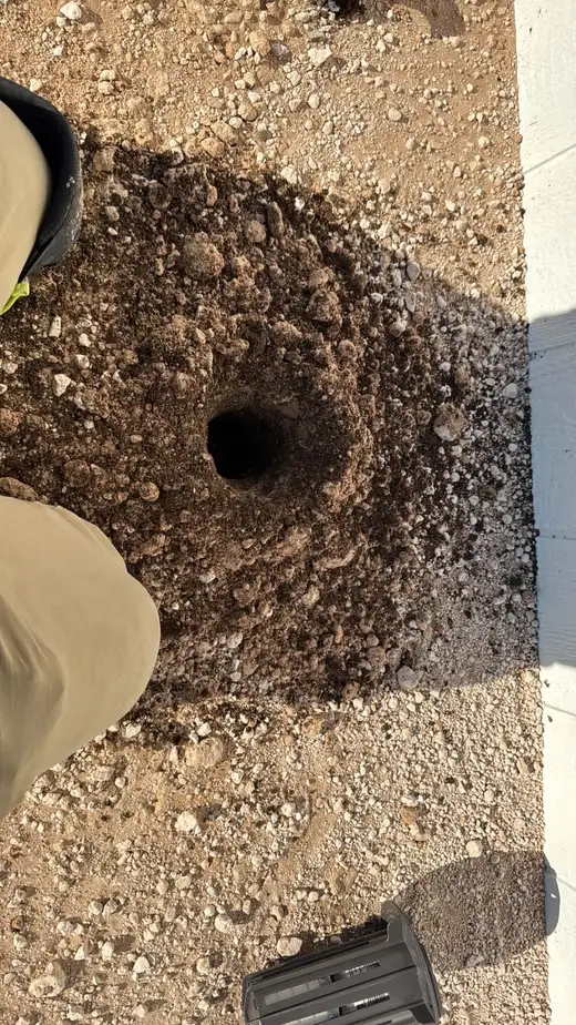 A freshly augered hole in caliche soil beside a slab, with a termite bait station ready to install